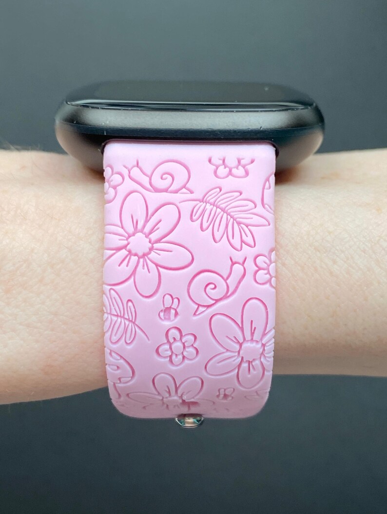 Puede incluir: Una correa de reloj de silicona rosa con un dise&ntilde;o floral y de caracol. La correa est&aacute; en la mu&ntilde;eca de una persona.