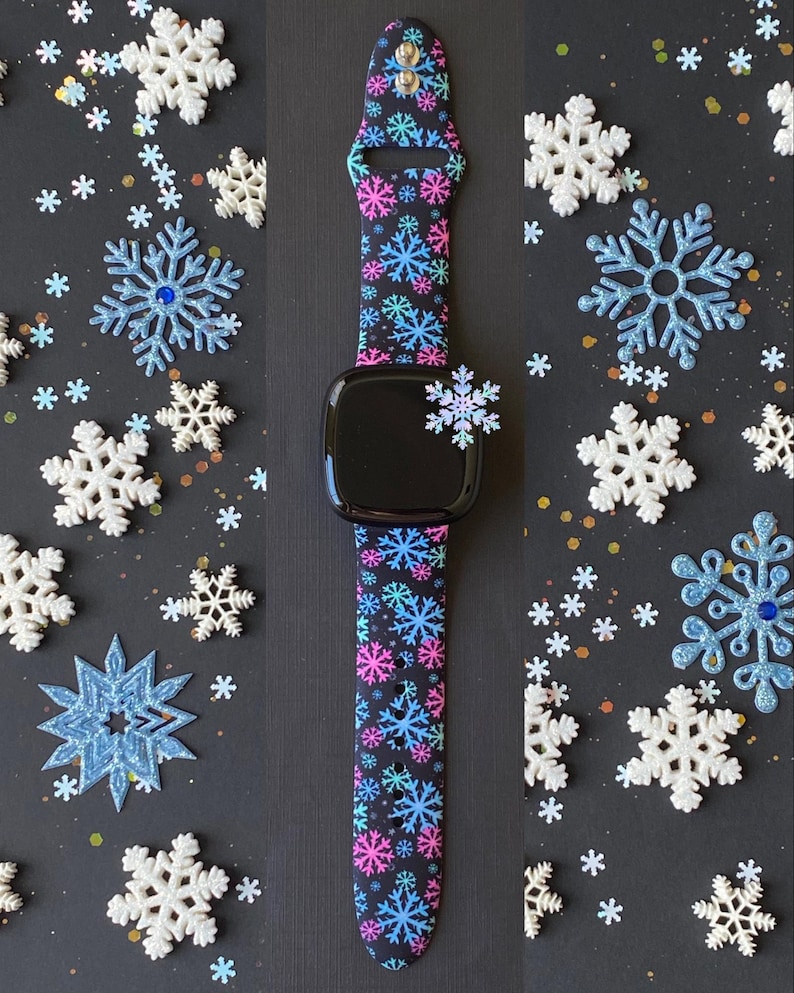 K&ouml;nnte beinhalten: Ein schwarzes Smartwatch-Armband mit einem Schneeflockenmuster in Pink, Blau und Wei&szlig;. Das Armband ist aus Silikon und kompatibel mit Apple Watch.