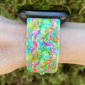 Puede incluir: Un reloj inteligente negro con una banda estampada de color azul brillante, verde y rosa con caballitos de mar. La banda tiene un fondo blanco con un patr&oacute;n repetitivo de caballitos de mar y coral.
