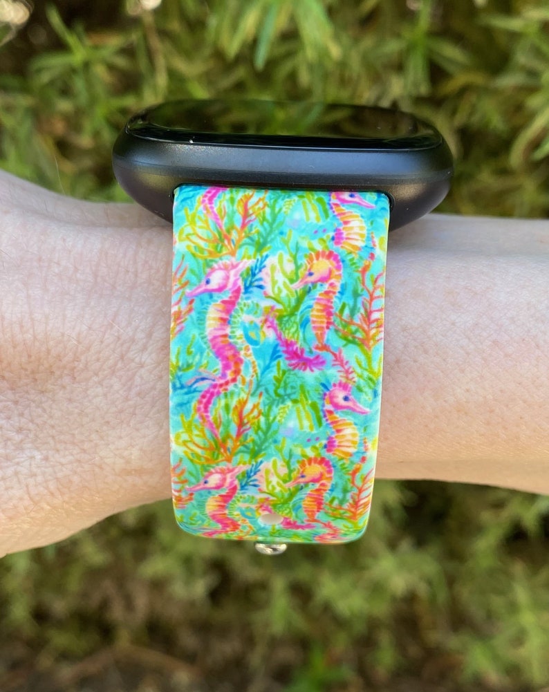 Puede incluir: Un reloj inteligente negro con una banda estampada de color azul brillante, verde y rosa con caballitos de mar. La banda tiene un fondo blanco con un patr&oacute;n repetitivo de caballitos de mar y coral.