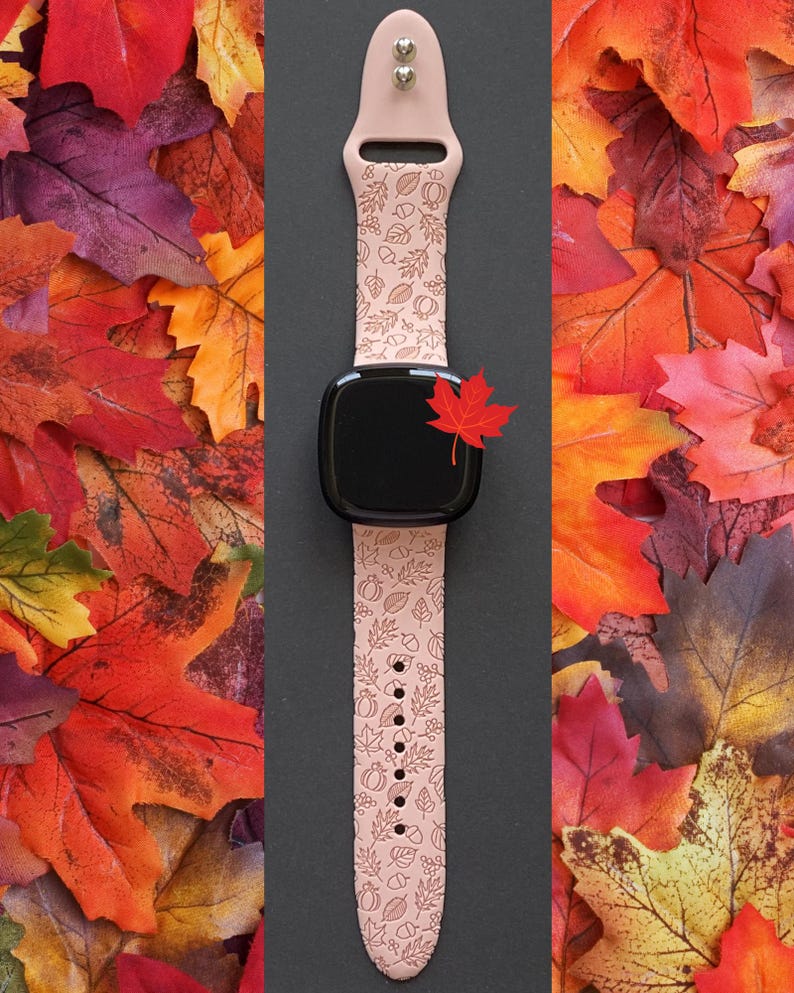 May include: A blush-colored smartwatch band with a fall-themed pattern of leaves, acorns, and pumpkins. The band is paired with a black smartwatch face and a small red maple leaf accent. The background features colorful autumn leaves.