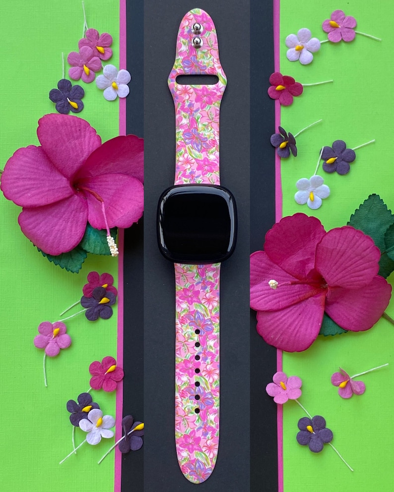 K&ouml;nnte beinhalten: Ein pinkfarbenes und wei&szlig;es Armband mit floralem Muster und einem schwarzen Zifferblatt. Das Armband besteht aus Silikon und hat einen Blumendruck. Das Zifferblatt ist rund und hat eine schwarze L&uuml;nette.
