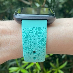 May include: A turquoise smartwatch band with a floral pattern. The band has a black watch face and is worn on a person's wrist.