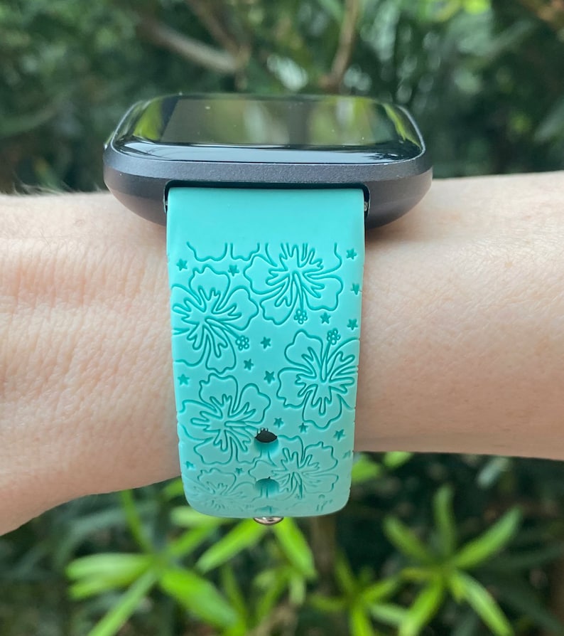 May include: A turquoise smartwatch band with a floral pattern. The band has a black watch face and is worn on a person's wrist.