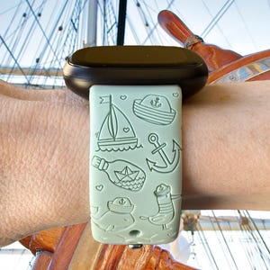 May include: A light green silicone watch band with a nautical theme. The band features a sailboat, anchor, bottle with a paper boat inside, whale, and a bird wearing a sailor hat.