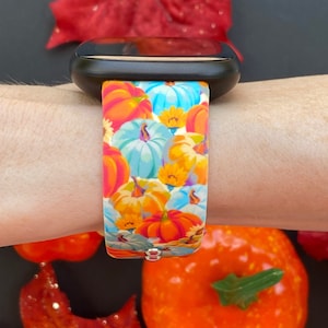 K&ouml;nnte beinhalten: Eine schwarze Smartwatch mit einem bunten Silikonband mit K&uuml;rbisdruck. Das Band zeigt eine Vielzahl von orangefarbenen, gelben, blauen und wei&szlig;en K&uuml;rbissen.