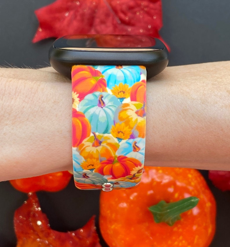 K&ouml;nnte beinhalten: Eine schwarze Smartwatch mit einem bunten Silikonband mit K&uuml;rbisdruck. Das Band zeigt eine Vielzahl von orangefarbenen, gelben, blauen und wei&szlig;en K&uuml;rbissen.