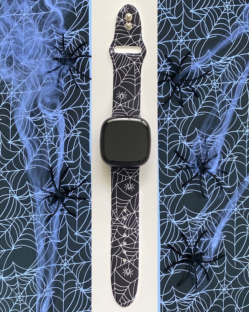 May include: A black smartwatch with a white spiderweb pattern band. The band has small spider silhouettes on it. The watch is on a white background with a black and blue spiderweb pattern on either side.