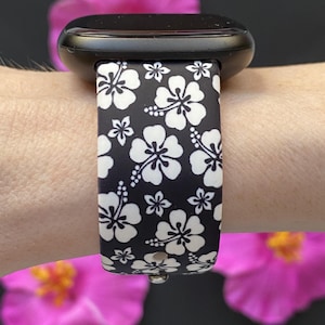 May include: Black smartwatch band with a white hibiscus flower pattern. The band is attached to a black smartwatch. The background features pink flowers.