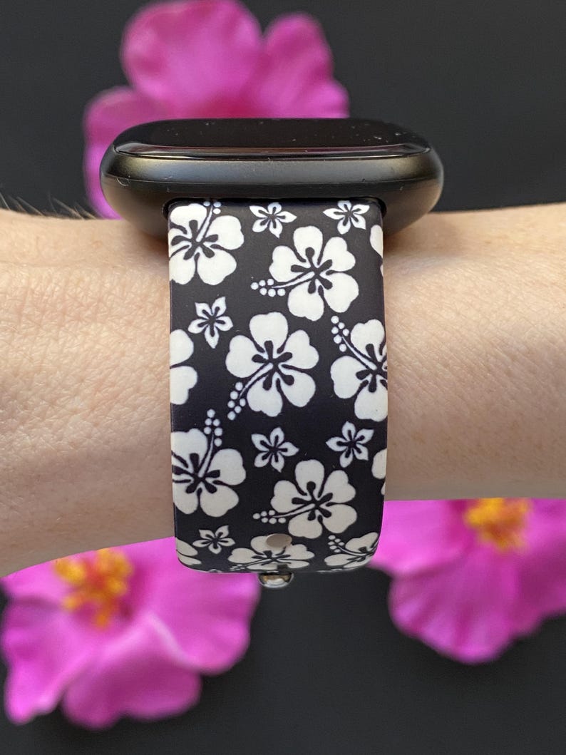 May include: Black smartwatch band with a white hibiscus flower pattern. The band is attached to a black smartwatch. The background features pink flowers.