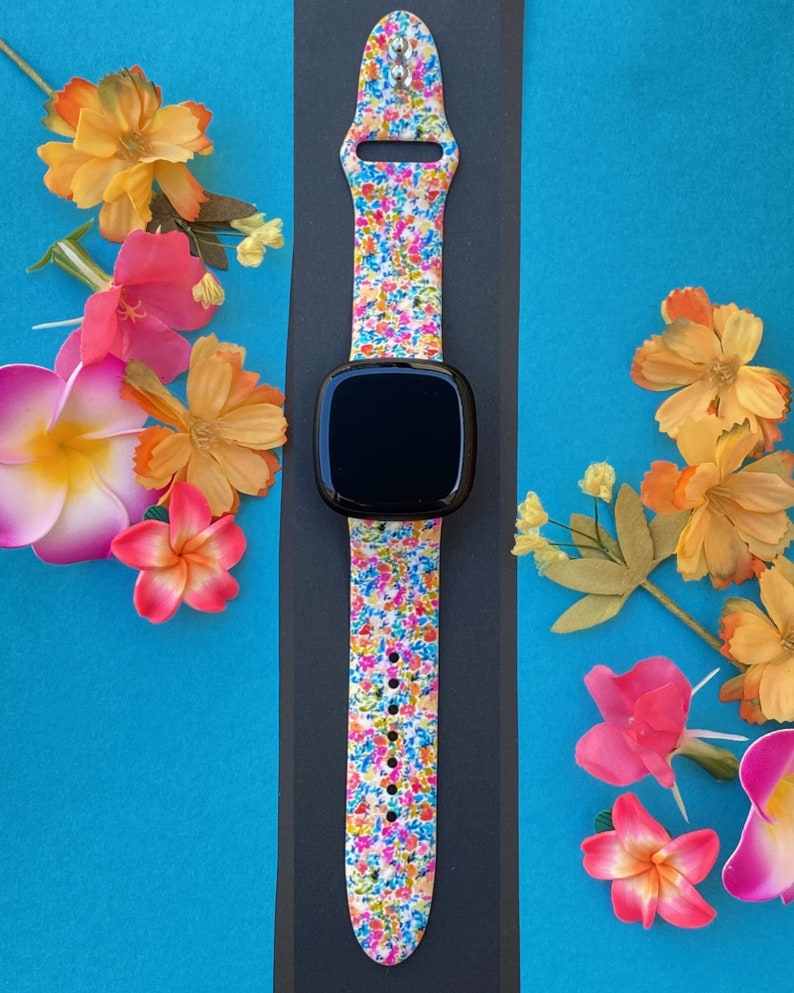 K&ouml;nnte beinhalten: Ein farbenfrohes Armband mit Blumenmuster und einem schwarzen Zifferblatt. Das Armband ist aus Silikon und hat einen wei&szlig;en Hintergrund mit kleinen rosa, blauen, gelben und orangefarbenen Blumen.