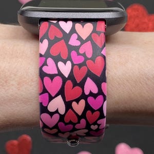 K&ouml;nnte beinhalten: Eine schwarze Smartwatch mit einem Armband, das mit rosa, roten und pinkfarbenen Herzen auf schwarzem Hintergrund gemustert ist. Die Uhr befindet sich an einem Handgelenk, mit herzf&ouml;rmigen Dekorationen im Hintergrund.
