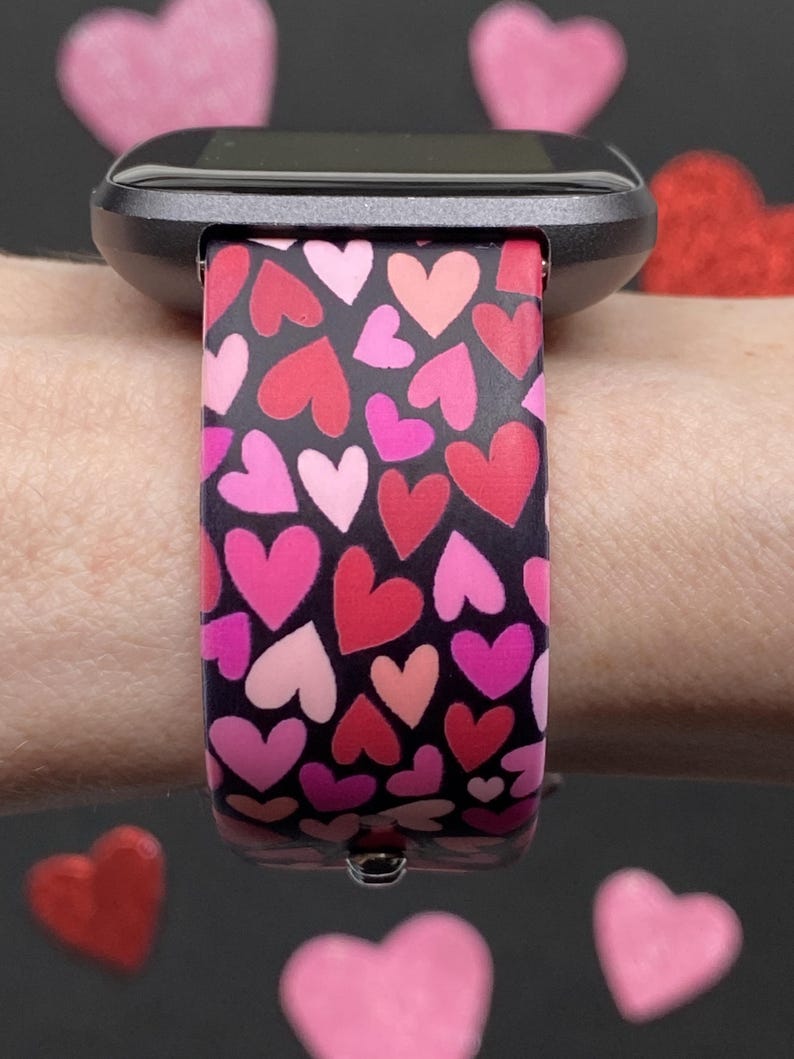 K&ouml;nnte beinhalten: Eine schwarze Smartwatch mit einem Armband, das mit rosa, roten und pinkfarbenen Herzen auf schwarzem Hintergrund gemustert ist. Die Uhr befindet sich an einem Handgelenk, mit herzf&ouml;rmigen Dekorationen im Hintergrund.