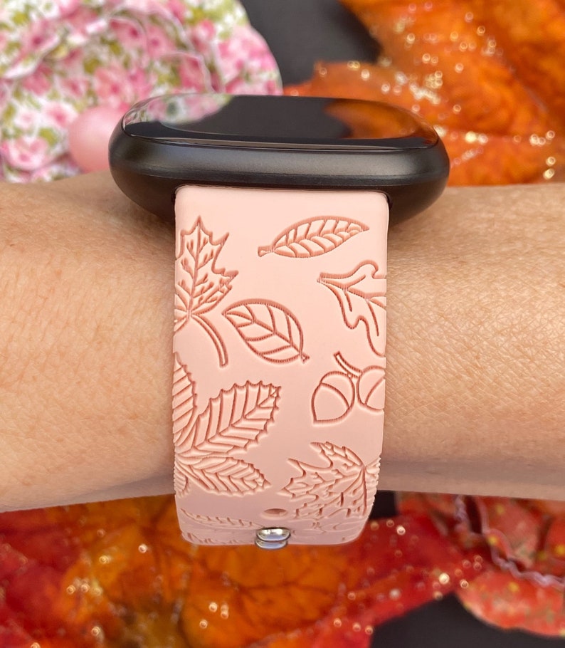 May include: A pink silicone watch band with a fall leaf design. The band has a black watch face and a silver buckle.