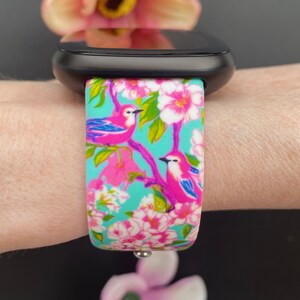K&ouml;nnte beinhalten: Ein t&uuml;rkisfarbenes Armband mit einem Blumen- und Vogelmuster. Das Armband zeigt rosa Bl&uuml;ten, gr&uuml;ne Bl&auml;tter und rosa V&ouml;gel mit blauen Fl&uuml;geln.