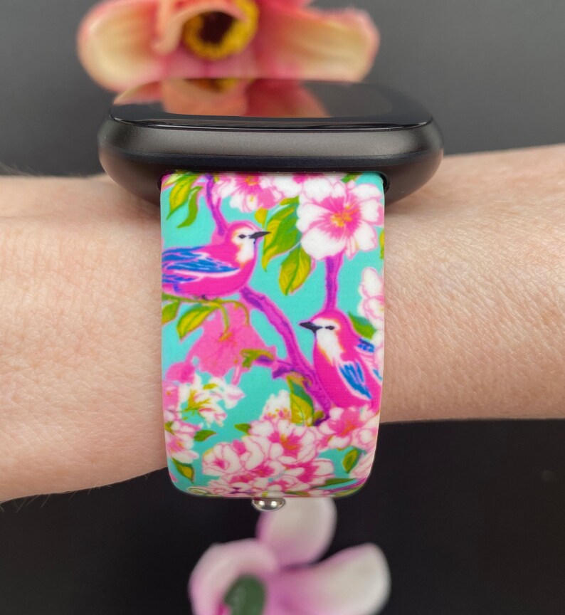 K&ouml;nnte beinhalten: Ein t&uuml;rkisfarbenes Armband mit einem Blumen- und Vogelmuster. Das Armband zeigt rosa Bl&uuml;ten, gr&uuml;ne Bl&auml;tter und rosa V&ouml;gel mit blauen Fl&uuml;geln.