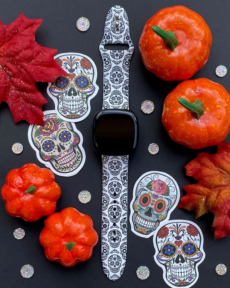 May include: A white and black watch band with a sugar skull pattern. The band is on a black surface with several small, sparkly, silver decorations. There are also several orange pumpkins and fall leaves on the surface.