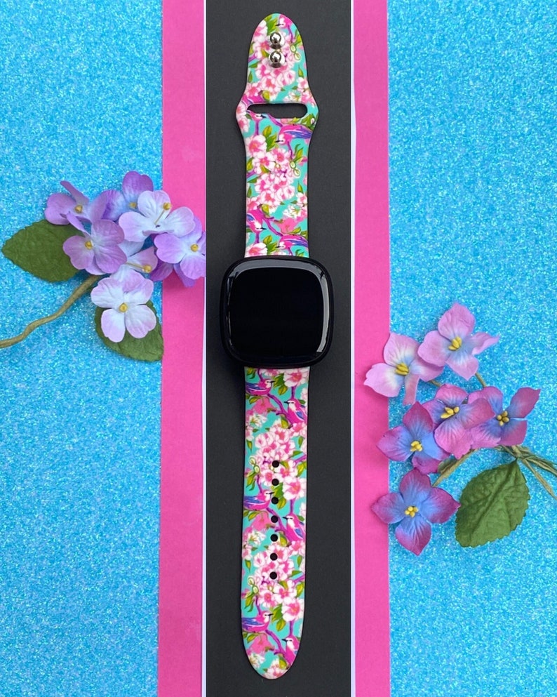 K&ouml;nnte beinhalten: Eine schwarze Smartwatch mit einem leuchtend pink- und blaufarbenen, floral gemusterten Armband mit pinkfarbenen Flamingos. Das Armband hat eine silberne Schlie&szlig;e.