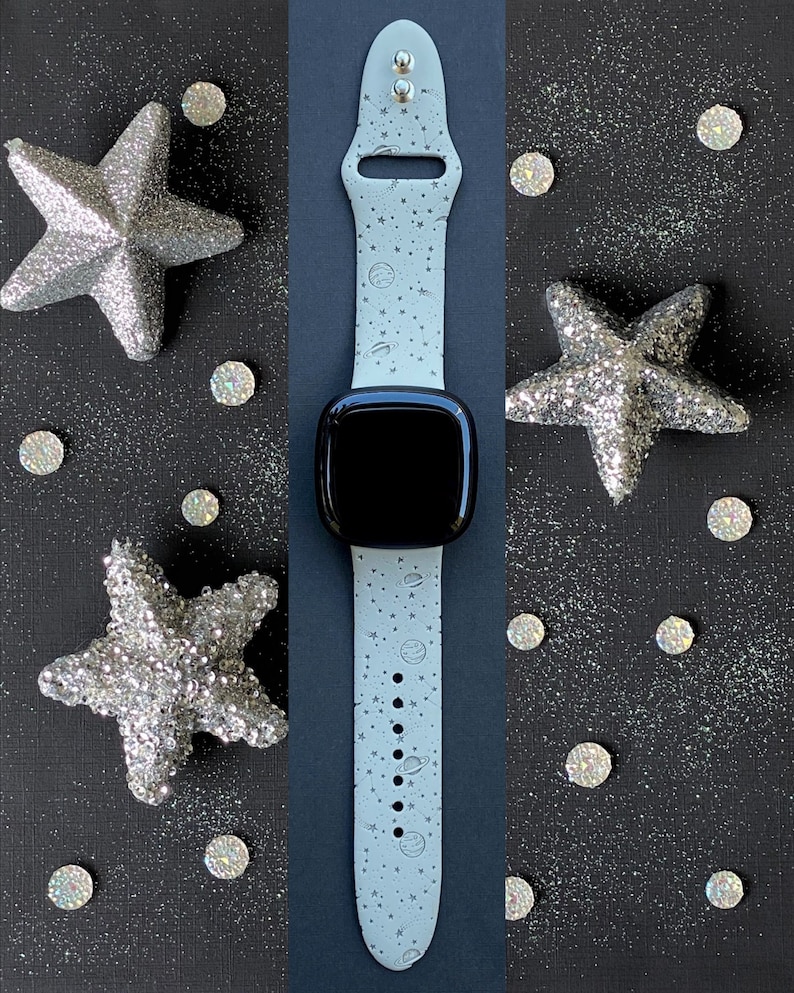 May include: A white Apple Watch band with a space-themed design featuring stars, planets, and a galaxy print. The band is shown on a black background with silver glitter.