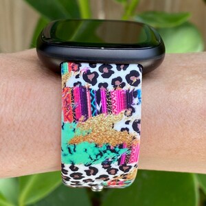 K&ouml;nnte beinhalten: Eine schwarze Smartwatch mit einem bunten, gemusterten Armband. Das Armband zeigt einen Leopardenprint, geometrische Muster und Farbschleier in Pink, Gr&uuml;n und Gold.