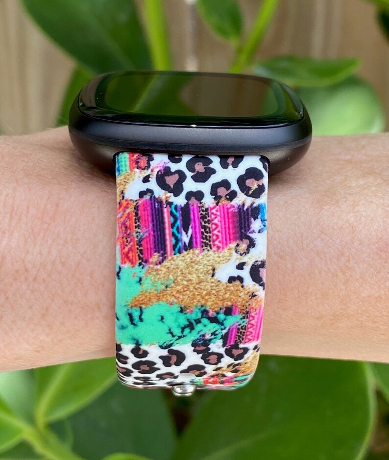 K&ouml;nnte beinhalten: Eine schwarze Smartwatch mit einem bunten, gemusterten Armband. Das Armband zeigt einen Leopardenprint, geometrische Muster und Farbschleier in Pink, Gr&uuml;n und Gold.