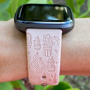 K&ouml;nnte beinhalten: Eine rosafarbene Smartwatch mit schwarzem Zifferblatt und einem Armband mit Kaktus-Motiv. Das Armband hat ein wei&szlig;es Kaktus-Design auf rosa Hintergrund.