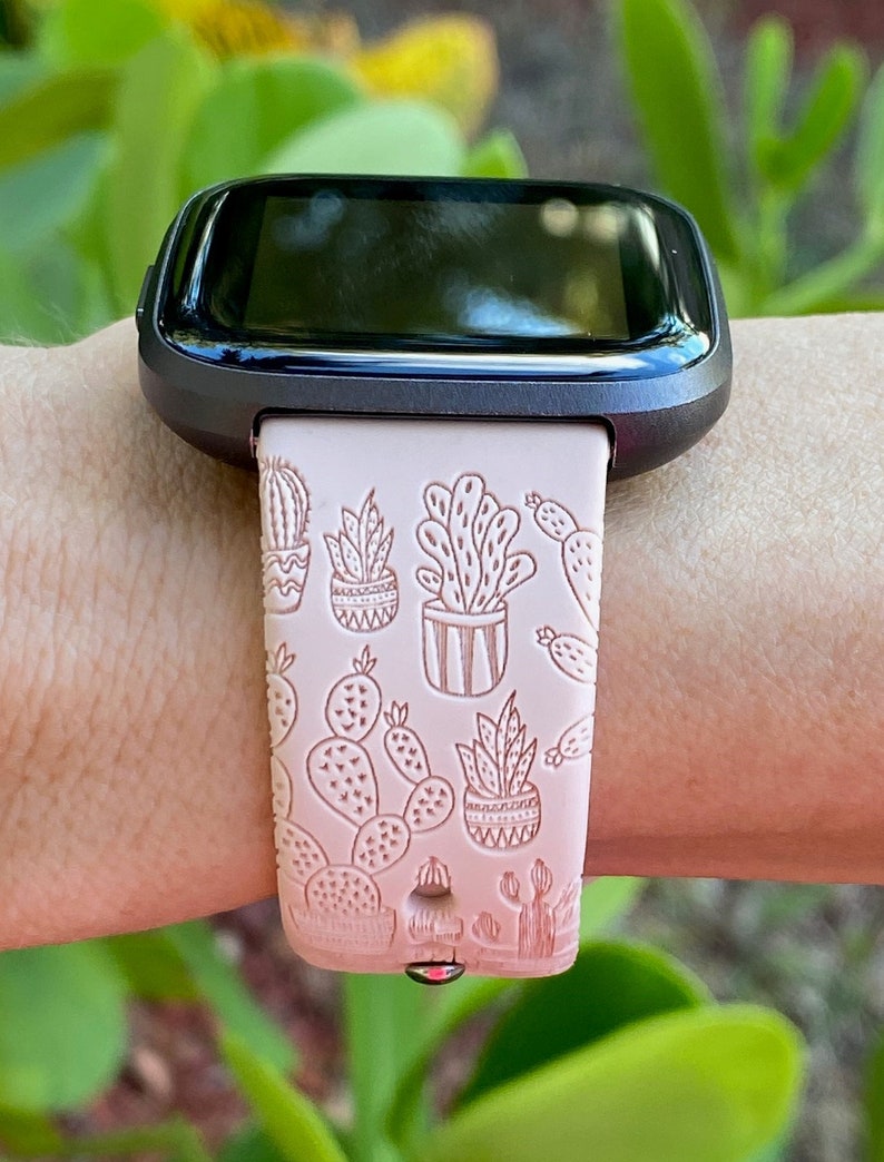 K&ouml;nnte beinhalten: Eine rosafarbene Smartwatch mit schwarzem Zifferblatt und einem Armband mit Kaktus-Motiv. Das Armband hat ein wei&szlig;es Kaktus-Design auf rosa Hintergrund.