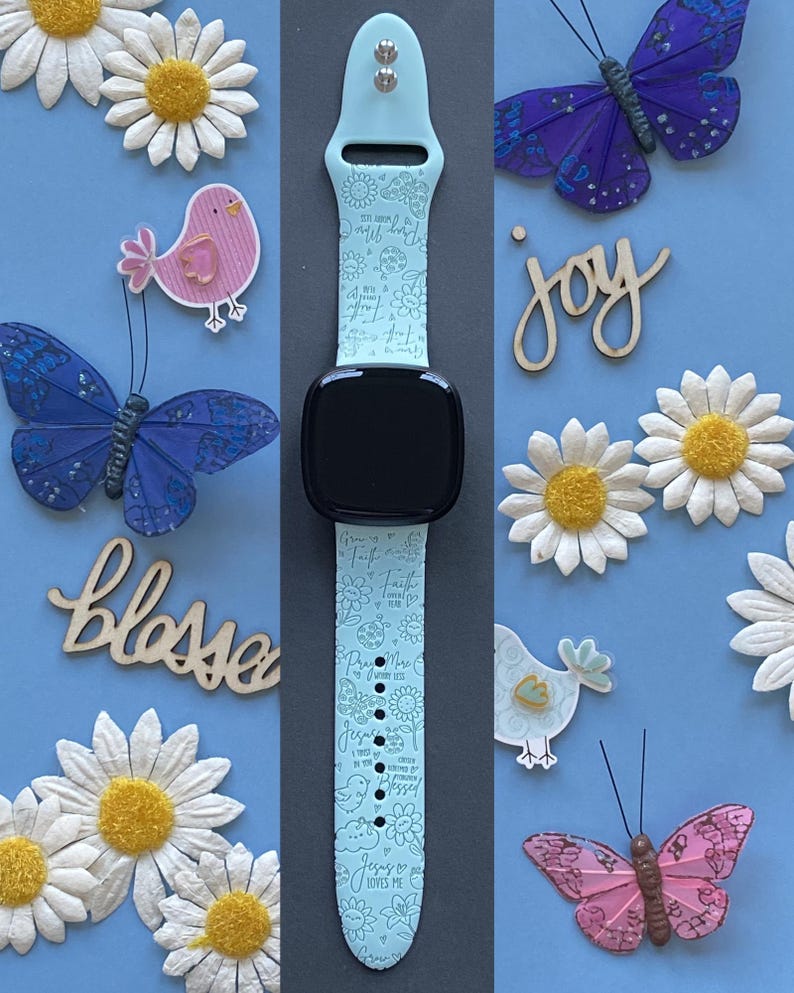 May include: A light blue smartwatch band with a black watch face, adorned with floral and script designs. The band is surrounded by decorative elements, including paper daisies, butterflies, and wooden cutouts spelling "joy" and "bless".