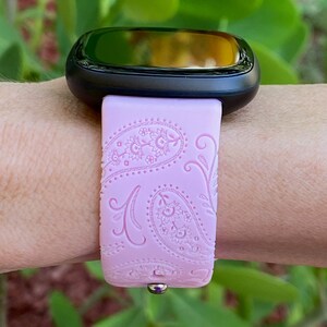 May include: A pink silicone watch band with a paisley pattern. The band is designed for a smartwatch with a black face.
