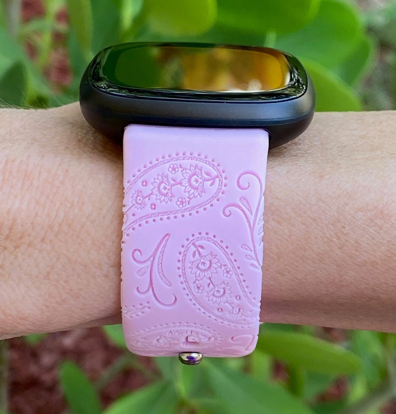 May include: A pink silicone watch band with a paisley pattern. The band is designed for a smartwatch with a black face.