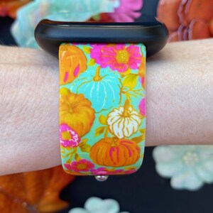 May include: A black Apple Watch with a colorful floral print band featuring pumpkins, flowers, and leaves. The band is a light blue background with orange, pink, and white accents.