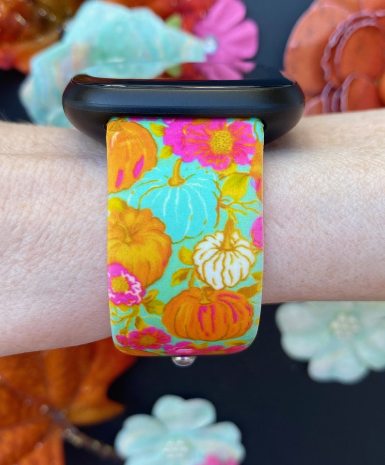 May include: A black Apple Watch with a colorful floral print band featuring pumpkins, flowers, and leaves. The band is a light blue background with orange, pink, and white accents.