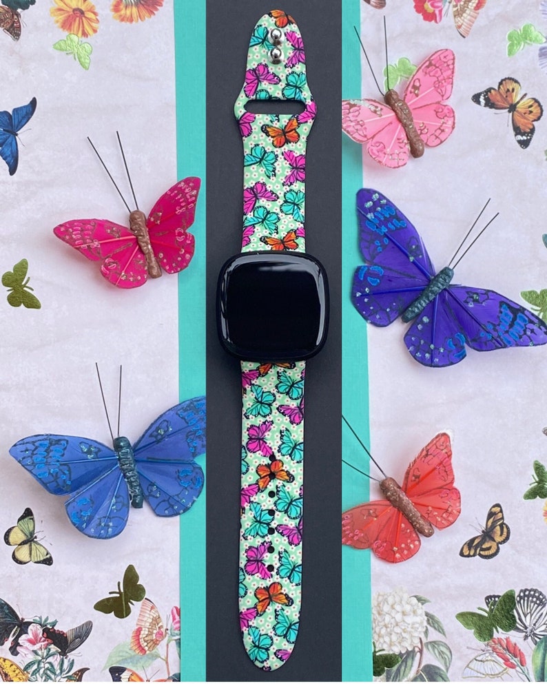 May include: A black smartwatch with a green, white, and pink butterfly patterned band. The band has a white background with small white dots and colorful butterflies.