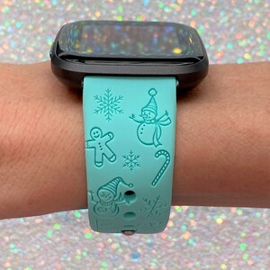 May include: A turquoise blue smartwatch band with a winter holiday theme. The band features a snowman, gingerbread man, candy cane, and snowflakes.