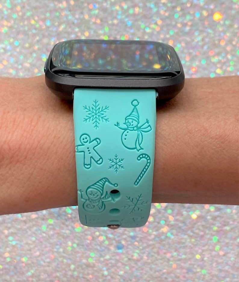 May include: A turquoise blue smartwatch band with a winter holiday theme. The band features a snowman, gingerbread man, candy cane, and snowflakes.
