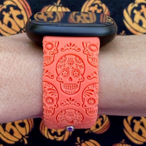 May include: A bright orange silicone watch strap with a sugar skull design. The strap is on a black smartwatch with a black screen.