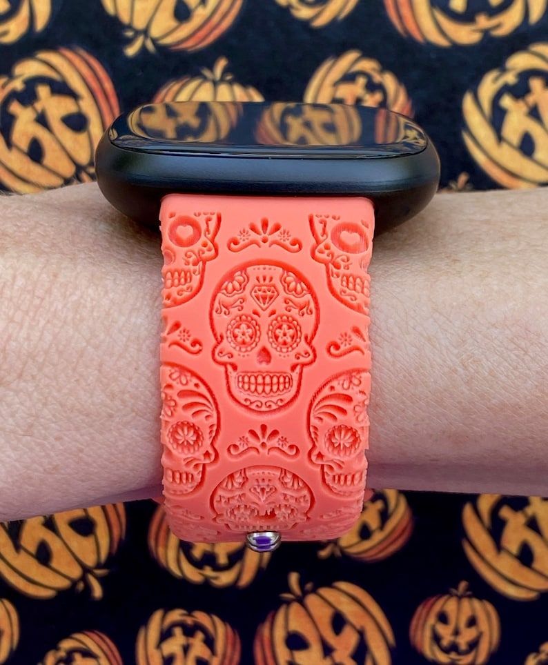 May include: A bright orange silicone watch strap with a sugar skull design. The strap is on a black smartwatch with a black screen.