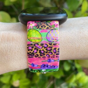 May include: A colorful watch band with a pink, green, and purple Easter egg design. The band has a leopard print pattern and floral accents.