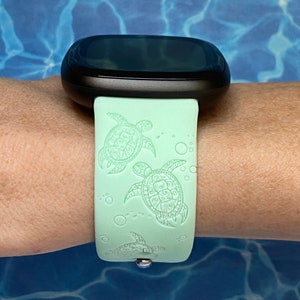 May include: A light green smartwatch band with a black watch face. The band has a repeating pattern of sea turtles and bubbles.