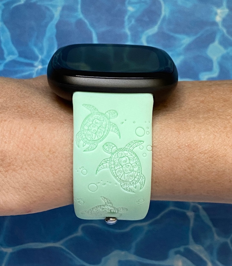May include: A light green smartwatch band with a black watch face. The band has a repeating pattern of sea turtles and bubbles.