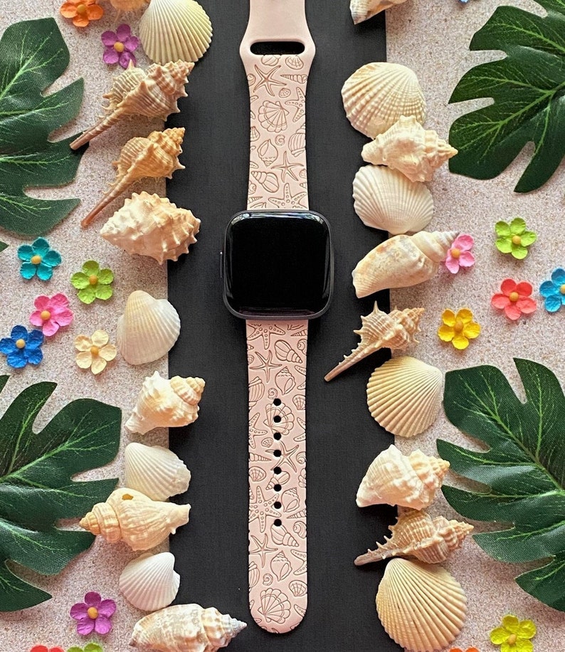 K&ouml;nnte beinhalten: Ein rosafarbenes Smartwatch-Armband mit einem Muschel- und Seesternmuster. Das Armband befindet sich auf einem schwarzen Hintergrund, umgeben von Muscheln und Blumen.