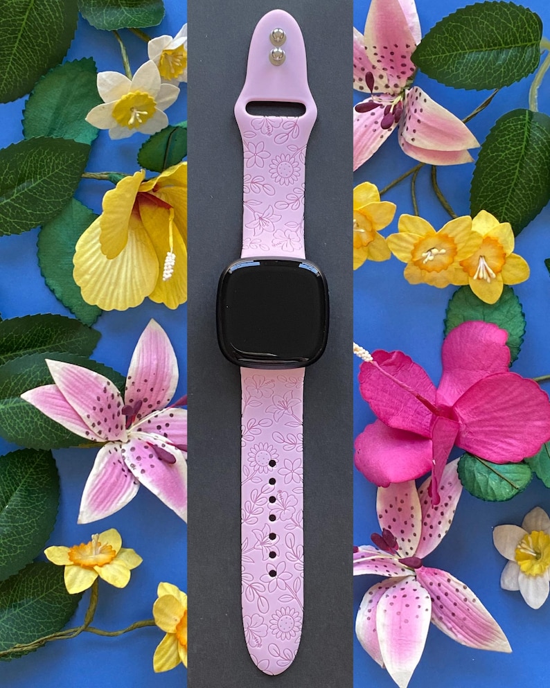 May include: A light purple smartwatch band with a floral pattern. The band is shown with a black smartwatch face. The background is a blue surface with yellow, pink, and white flowers and green leaves.
