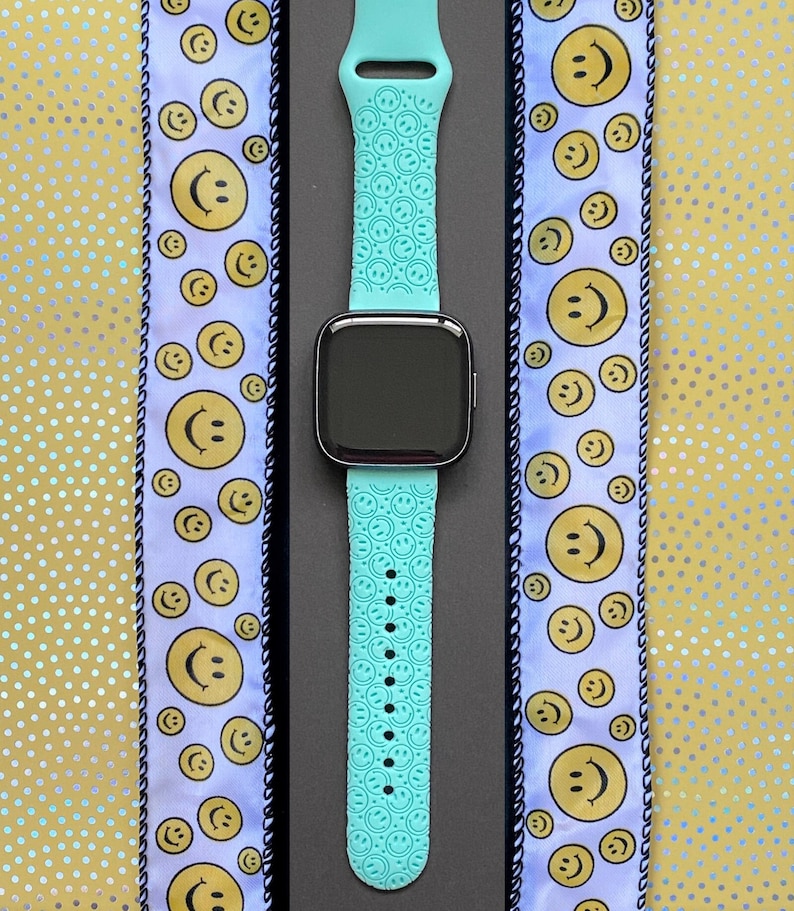 May include: A turquoise blue Apple Watch band with a pattern of smiling faces. The band is on a black background.