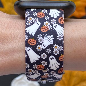 May include: A black smartwatch band with a Halloween-themed pattern. The band features white ghosts, orange pumpkins, and white flowers on a dark blue background. The band is attached to a black smartwatch.