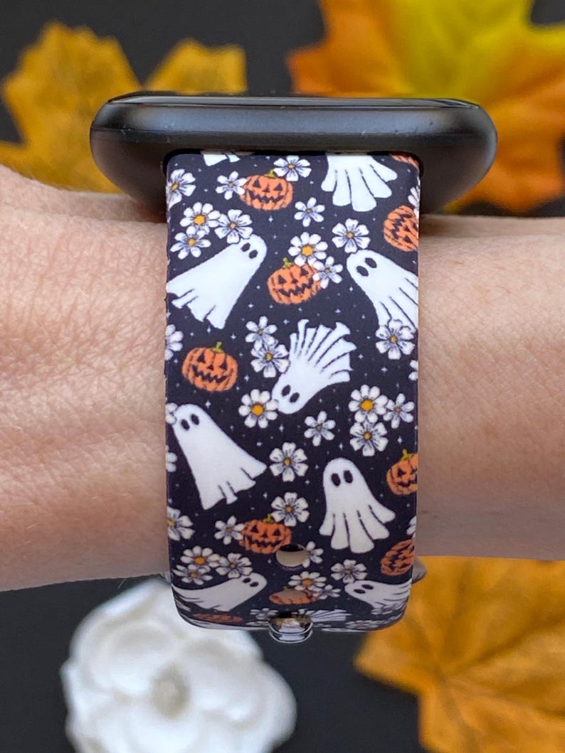 May include: A black smartwatch band with a Halloween-themed pattern. The band features white ghosts, orange pumpkins, and white flowers on a dark blue background. The band is attached to a black smartwatch.