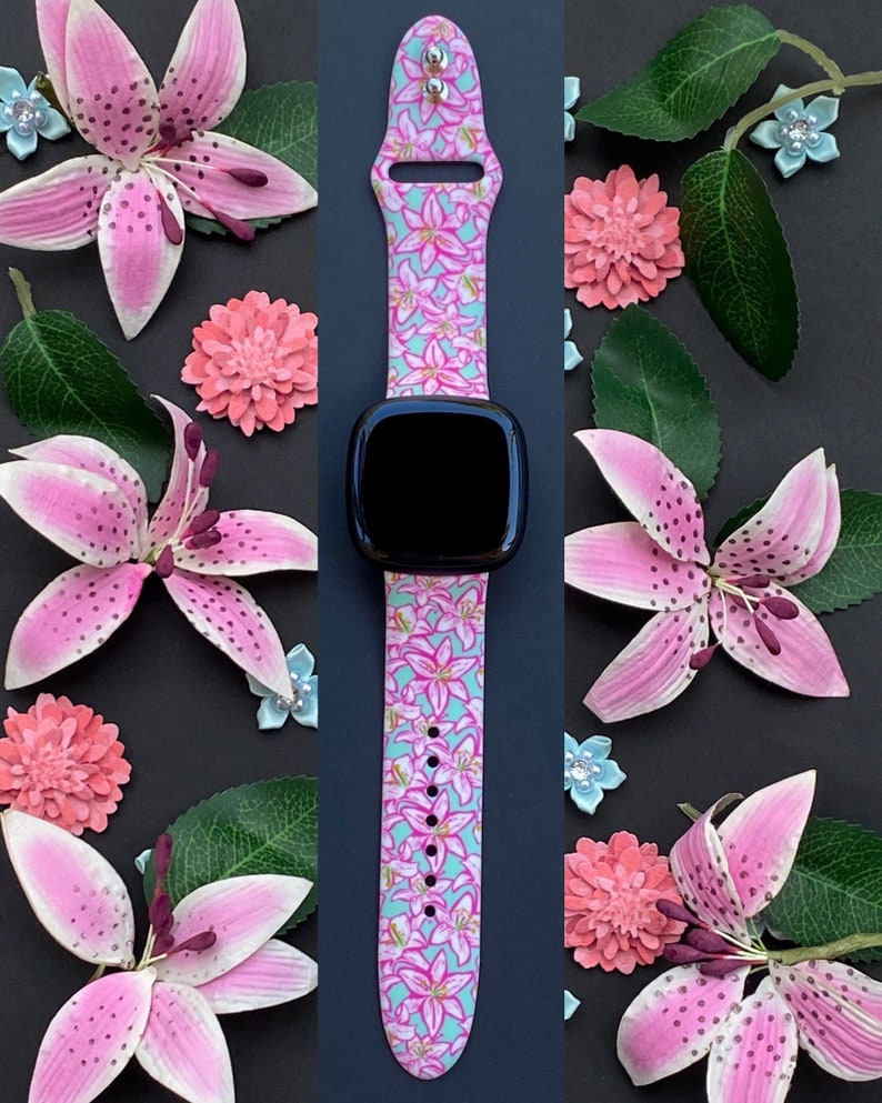 Puede incluir: Una correa de reloj con estampado floral rosa y azul con una esfera negra. La correa est&aacute; hecha de silicona y tiene un dise&ntilde;o floral con lirios rosas sobre un fondo azul claro.