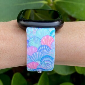 K&ouml;nnte beinhalten: Eine schwarze Smartwatch mit einem blauen und pinkfarbenen Armband mit Muschelmuster.
