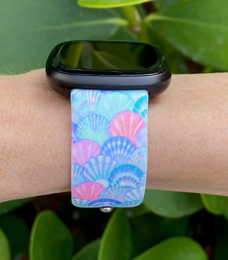 K&ouml;nnte beinhalten: Eine schwarze Smartwatch mit einem blauen und pinkfarbenen Armband mit Muschelmuster.