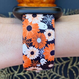 May include: A black watch strap with a Halloween-themed print. The strap features orange and white flowers, white ghosts, and black spiderwebs on a black background.
