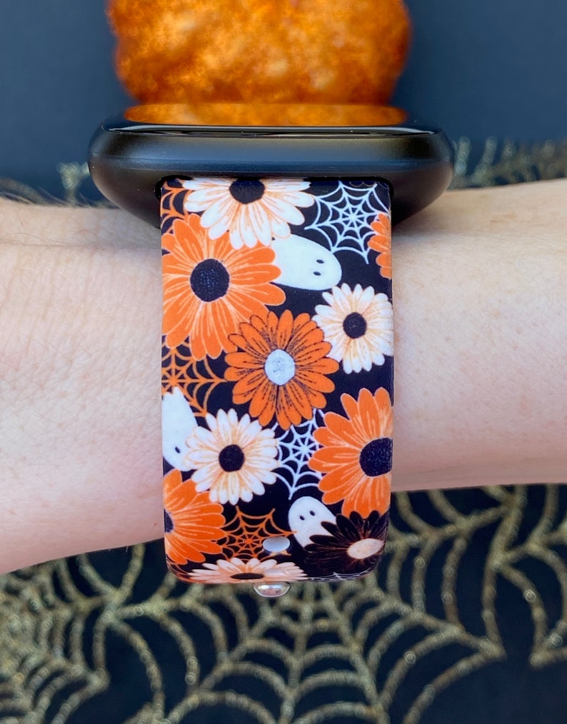 May include: A black watch strap with a Halloween-themed print. The strap features orange and white flowers, white ghosts, and black spiderwebs on a black background.